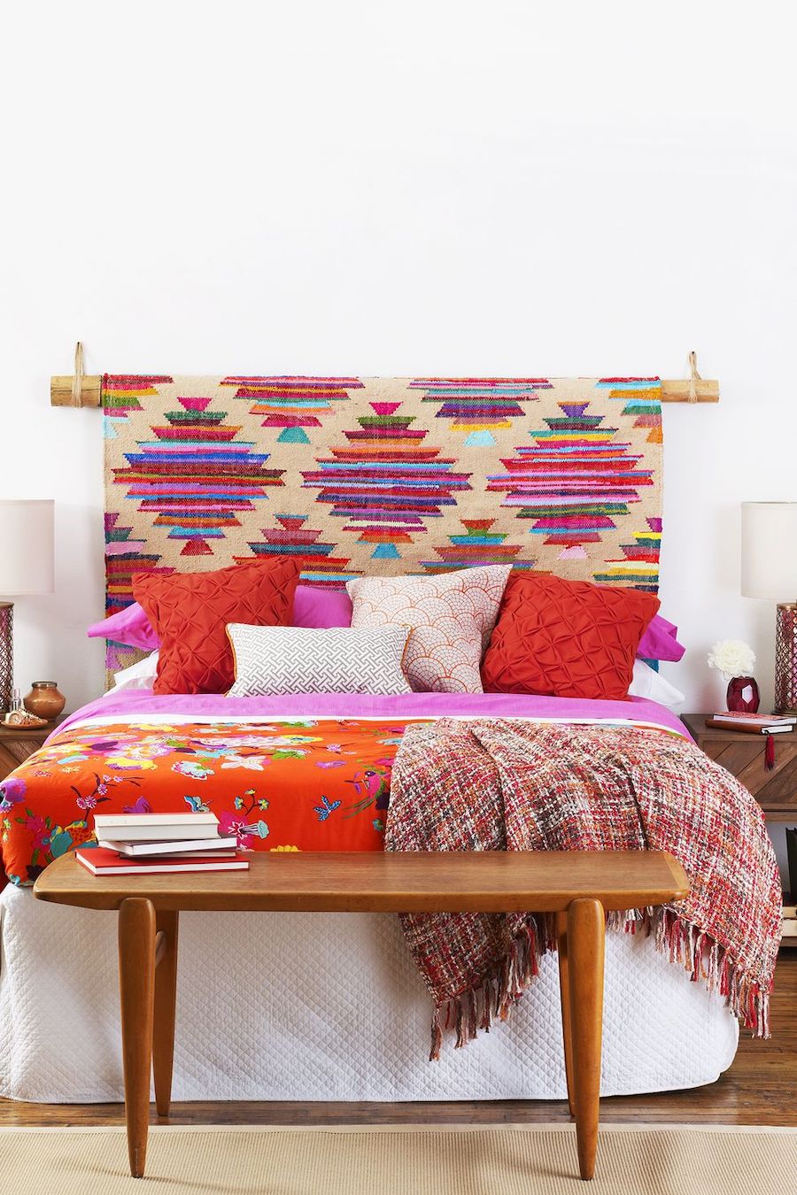 Dormitorio con pared blanca y piso de madera. En vez de respaldo de cama hay una alfombra de colores. Los cojines de la cama son rojos y rosados. Y también hay mantas en tonos rojizos. 
