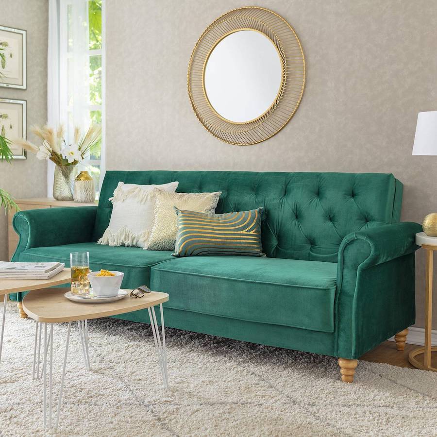 Sala de estar con un sofá cama verde esmeralda con tres cojines con detalles dorados sobre él. Está frente a dos mesas de centro con patas metálicas blancas y superficie de madera. En el muro hay un gran espejo redondo con marco dorado.