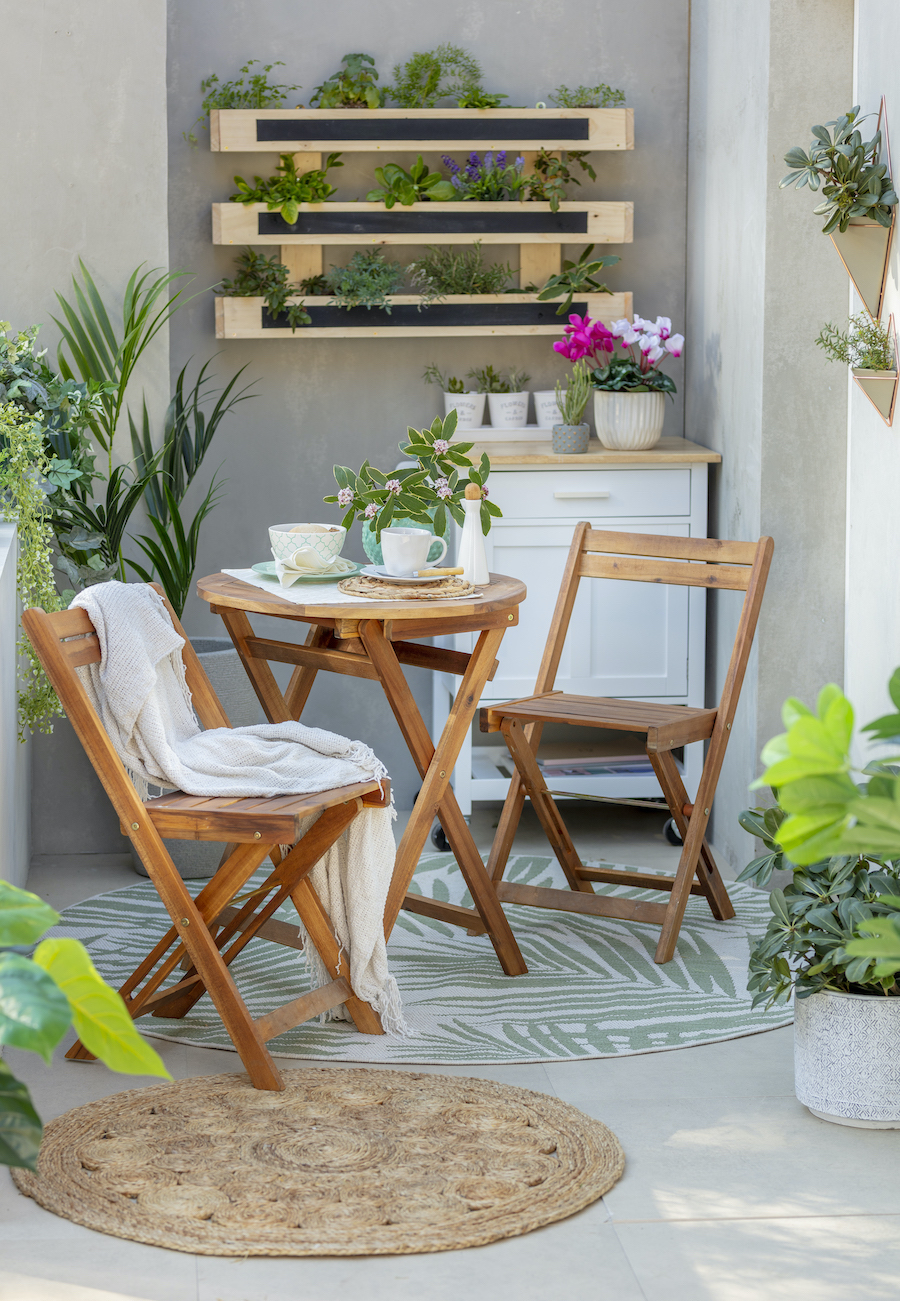 Terraza con mesa y sillas de madera, alfombra blanca con hojas verdes y una alfombra de yute redonda. Alrededor hay distintos tipos de plantas, tanto en el suelo como en el muro, en un huerto vertical y sobre una mesa tipo buffet.