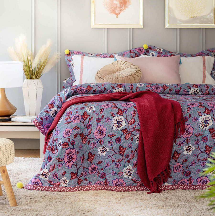 Dormitorio de paredes gris claro, piso de madera con una gran alfombra peluda blanca y una cama con un cobertor con diseño de flores y hojas en colores rosado, magenta, blanco y celeste. Sobre la cama hay una manta magenta y cojines en tonos pastel.