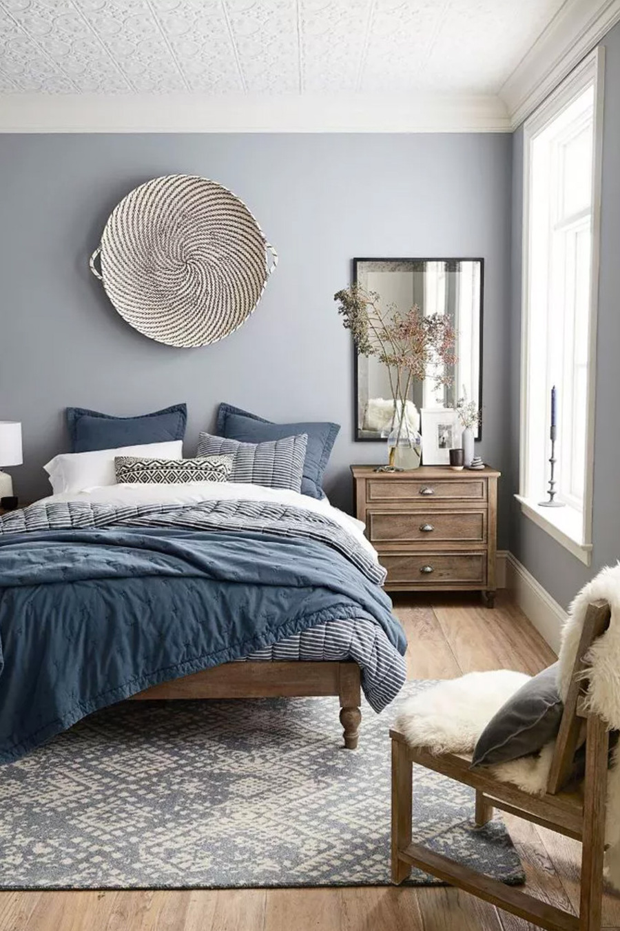 Dormitorio matrimonial con pared azul grisáceo claro, techo blanco y piso de madera clara. Hay una alfombra en tonos grises y crema, una cama de madera sin respaldo y con ropa de cama azulados y grises. Hay un velador de madera con tres cajones y un espejo sobre él, colgando de la pared. 