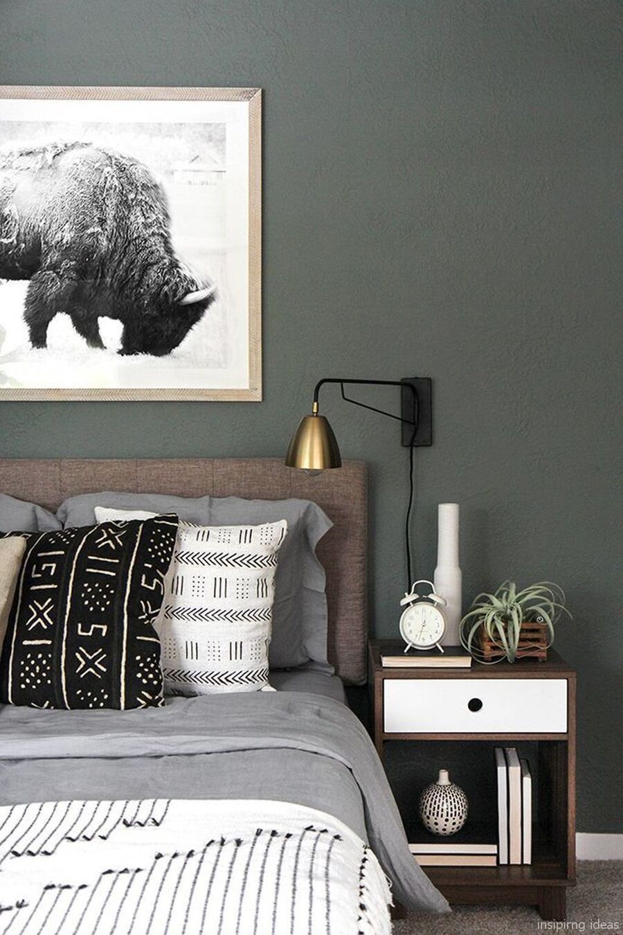 Detalle de un dormitorio con muro verde grisáceo oscuro y un cuadro con una fotografía en blanco y negro. Debajo está la cama con ropa de cama grises y cojines con diseños en blanco, negro y gris. Junto a la cama hay un velador de madera con cajón blanco.