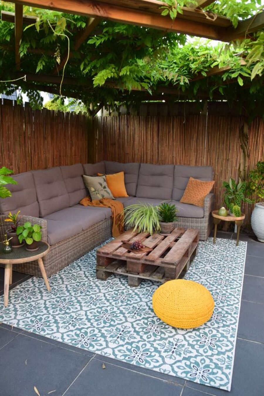 Terraza de un patio con árboles que dan sombra. El piso es gris oscuro y sobre él hay una alfombra blanca con diseño turquesa. Sobre ella hay una mesa de centro con pallets y un pouf amarillo y redondo. El sofá es seccional en forma de ele y tiene cojines grises y anaranjados.