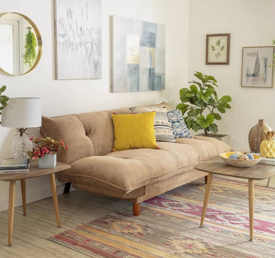 Sala de estar con un futón café acolchado. Sobre él hay tres cojines de distintos colores y diseños. La pared del fondo es blanca y en ella hay dos canvas con diseño moderno, y un espejo redondo con marco dorado. El piso es de madera y sobre él hay una alfombra en tonos rojizos. 