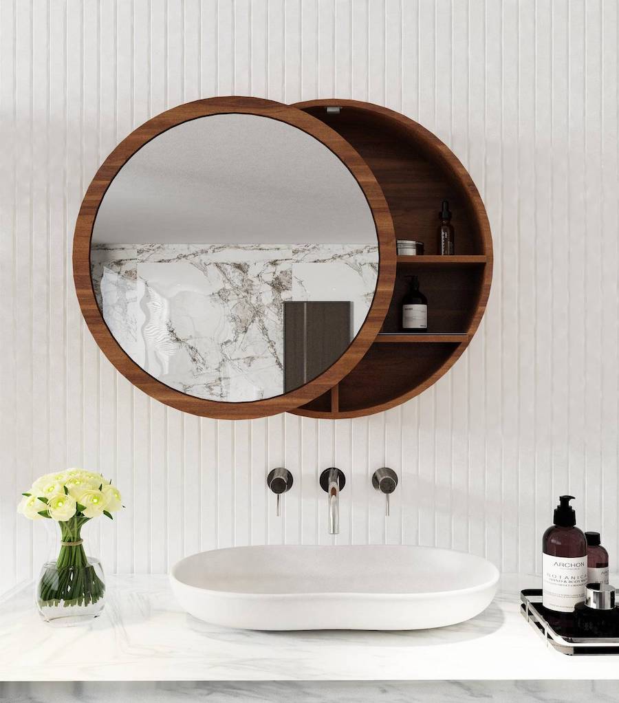 Baño con espejo tipo botiquín. Es redondo, de madera oscura y su espejo se corre para tener acceso a unas mini repisas en su interior. Está pegado a un muro con azulejos blancos y debajo hay un lavamanos blanco sobre una superficie de mármol.