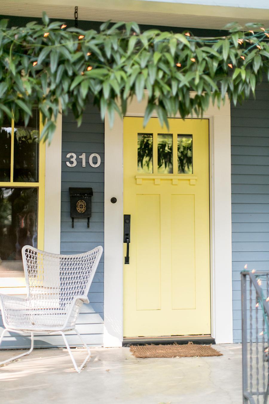 Puerta de entrada a una casa en color amarillo. Tiene manilla negra y el marco de la puerta es blanco. A su costado está el número de la casa, una ventana con marco blanco y detalles amarillos. El color de la casa es azul grisáceo. .