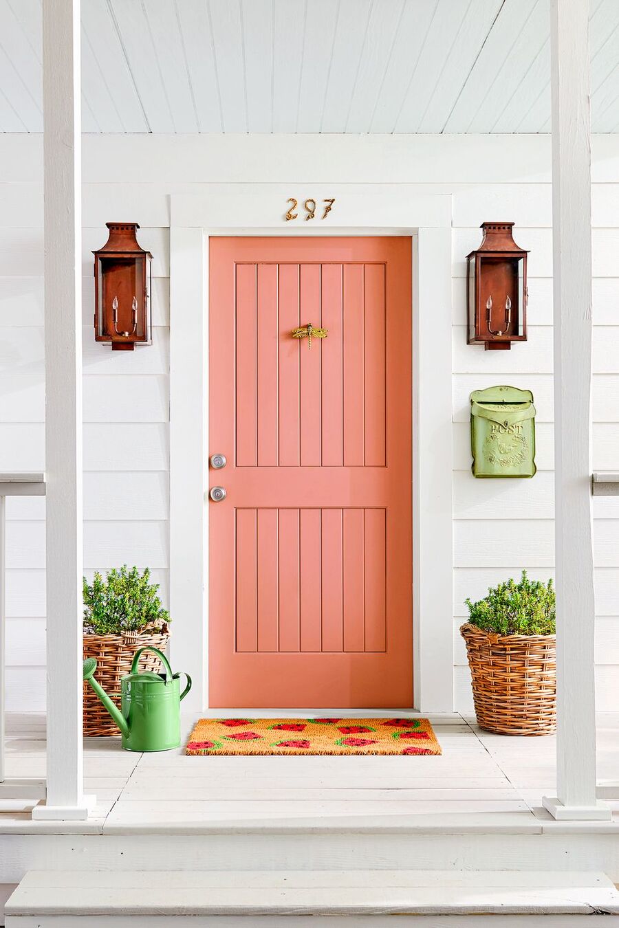 Puerta de entrada a una casa en color rosado. El techo, los muros y el piso son blancos. A cada costado de la puerta hay unos focos en color rojizo, dos canastos de mimbre con plantas, una regadera verde y un buzón verde claro. También hay un limpiapiés con diseño de sandías. 