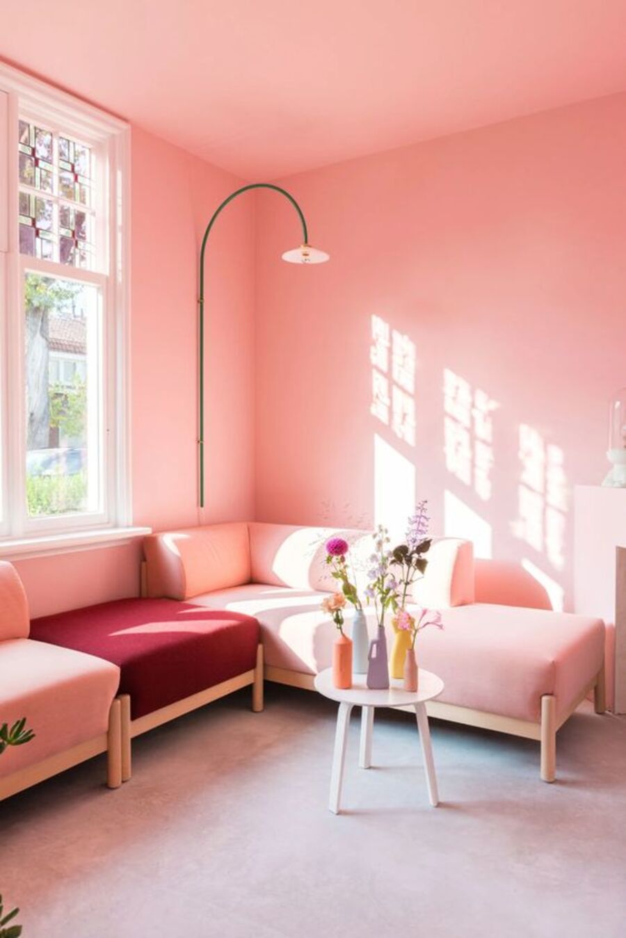 Sala de estar con techo y paredes rosado pastel. El sofá es del mismo color, pero uno de los cojines es burdeos. La mesa de centro tiene pequeños floreros de colores con flores en cada uno. 