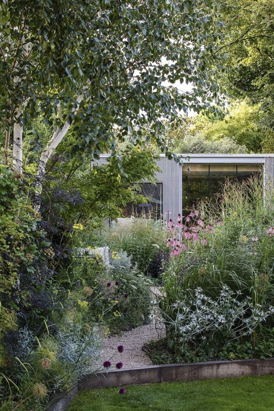 Jardín de una casa con muchos árboles, plantas y arbustos. Entre medio hay un camino en zigzag de gravilla blanca, que conecta la casa con el patio. 