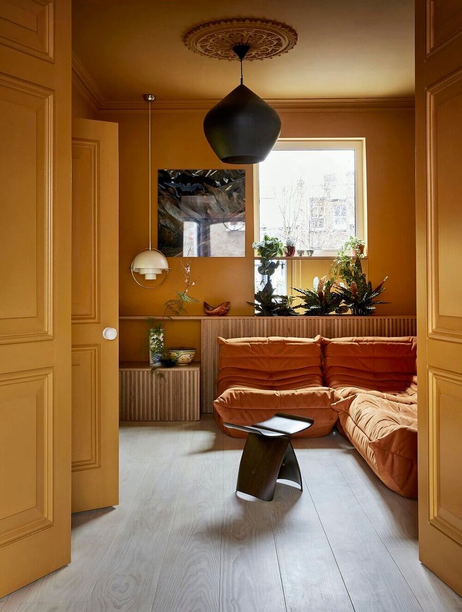 Living de puertas, techos y paredes de color mostaza, el piso es de madera grisácea y el sofá es anaranjado. El mueble del fondo es de madera y tiene diversas plantas sobre él. 