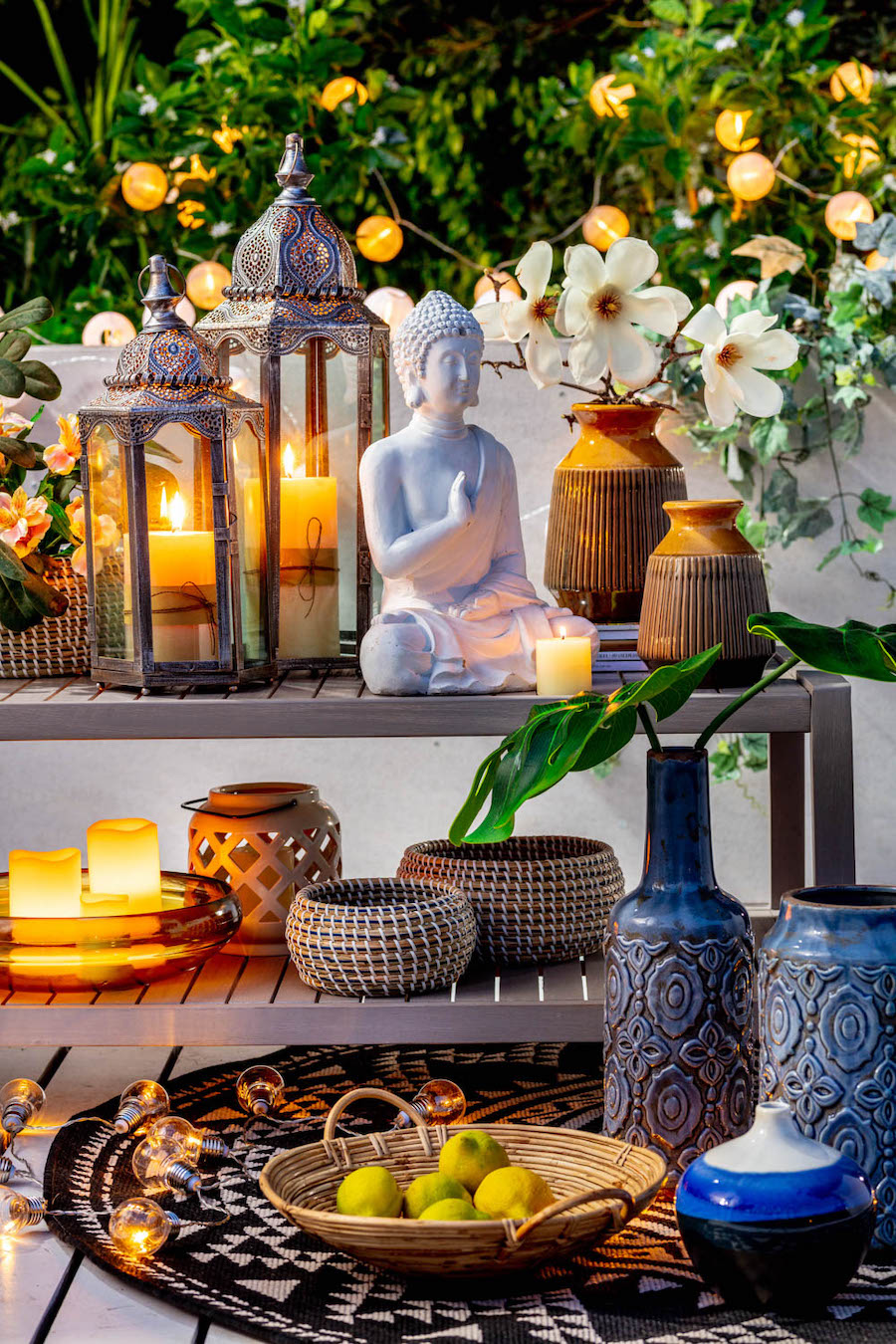 Detalle de un mueble tipo estante de dos niveles con distintos objetos decorativos, desde velas y fanales hasta figuras y floreros. Alrededor hay plantas, guirnaldas de luces y frascos decorativos.