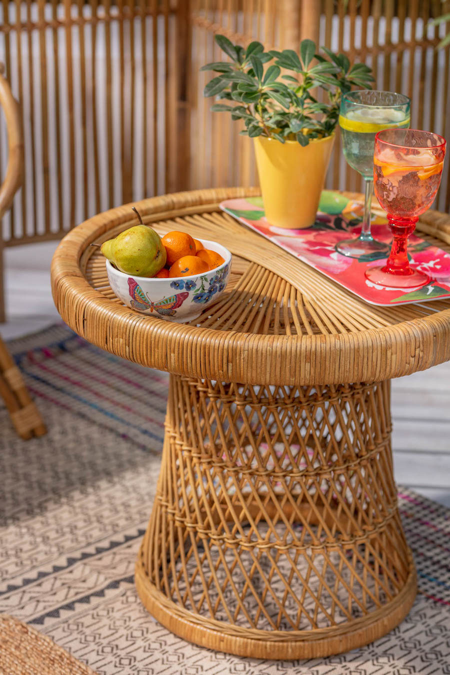 Detalle de una mesa lateral de mimbre redonda, con copas, una planta y un bowl con fruto sobre ella. 