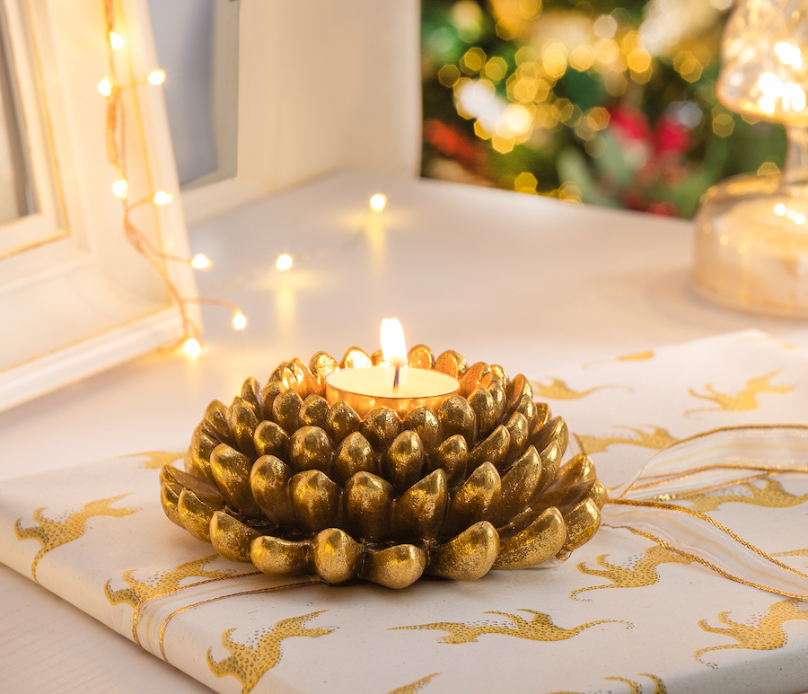 Detalle de un pequeño candelabro dorado con forma de piña y una vela encendida en su interior. Está sobre un paño blanco con diseño de tigres y alrededor hay luces navideñas.