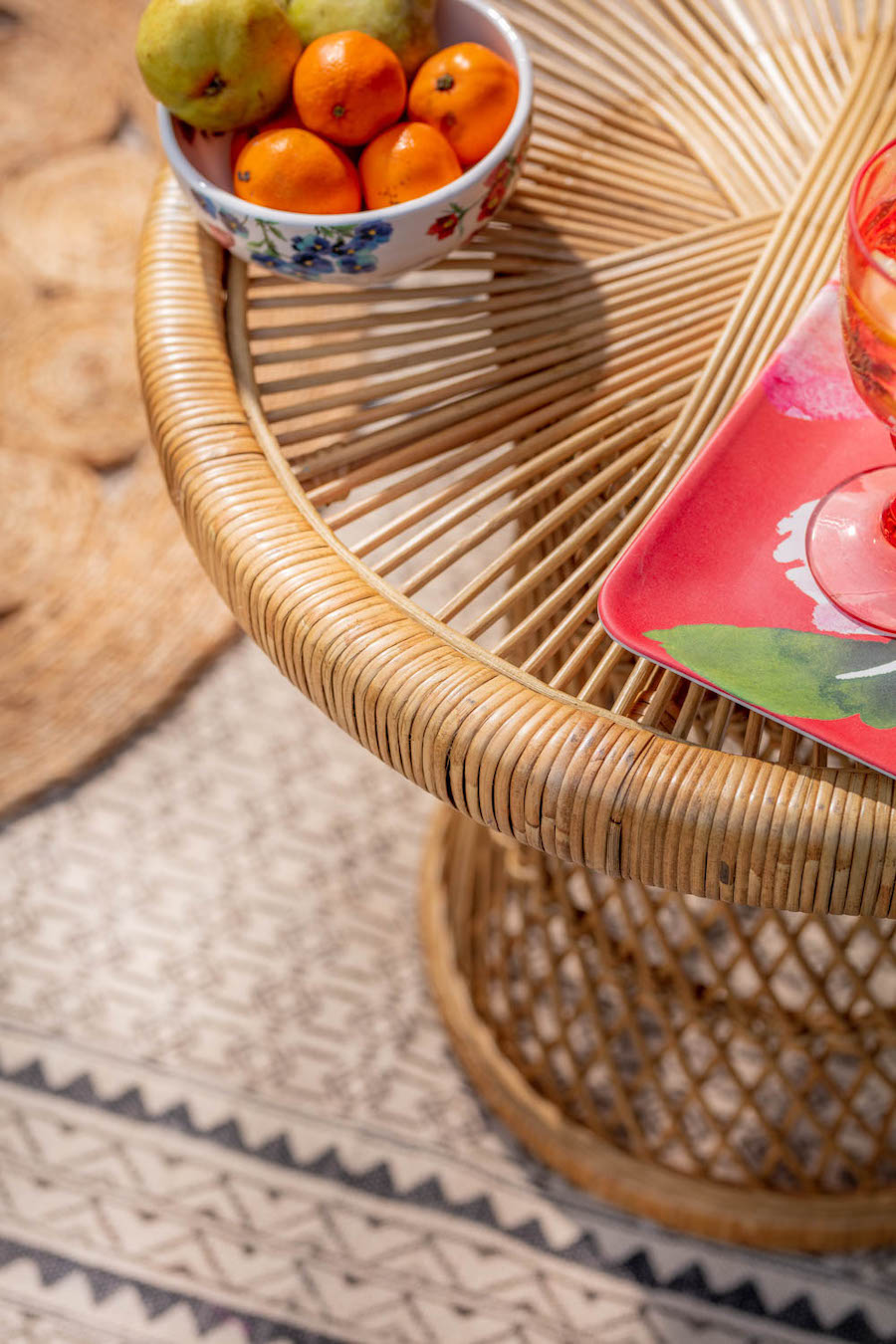Detalle de una mesa auxiliar de mimbre vista desde arriba. Sobre ella hay un bowl con frutas y una cosa anaranjada sobre una bandeja de melamina roja con diseño. 