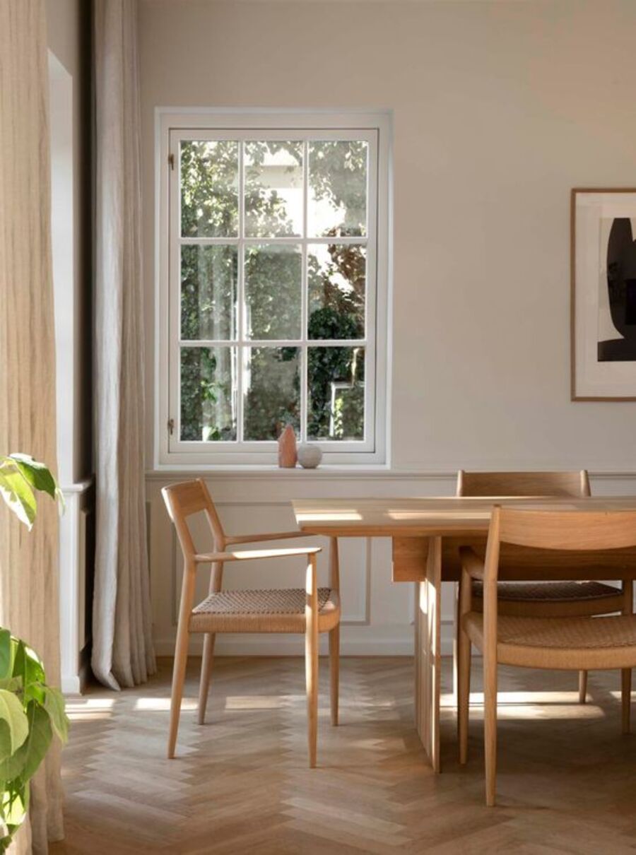 Comedor de piso de madera clara, mesa y sillas de madera y muros con molduras en la parte inferior. En uno de ellos hay una ventana con marco blanco que da hacia un jardín con vegetación.