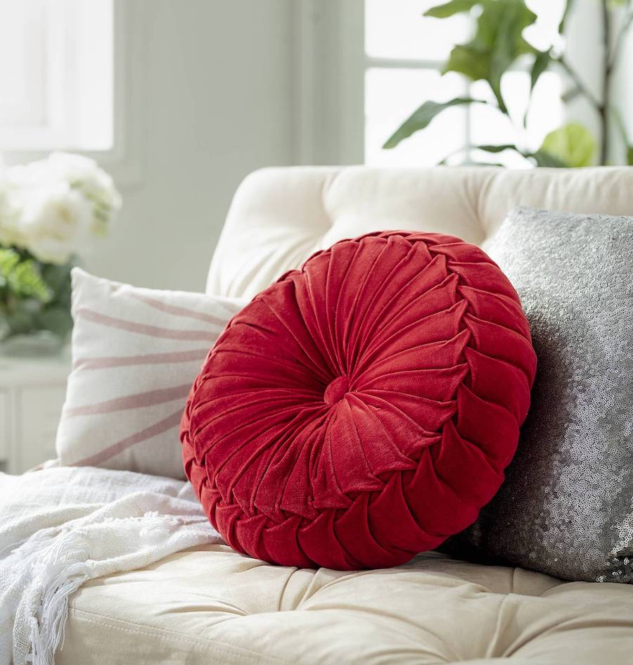 Detalle de un sofá blanco con un cojín plateado, uno blanco con líneas rosadas y uno de forma circular en color magenta. De fondo hay plantas y un muro con ventanas.