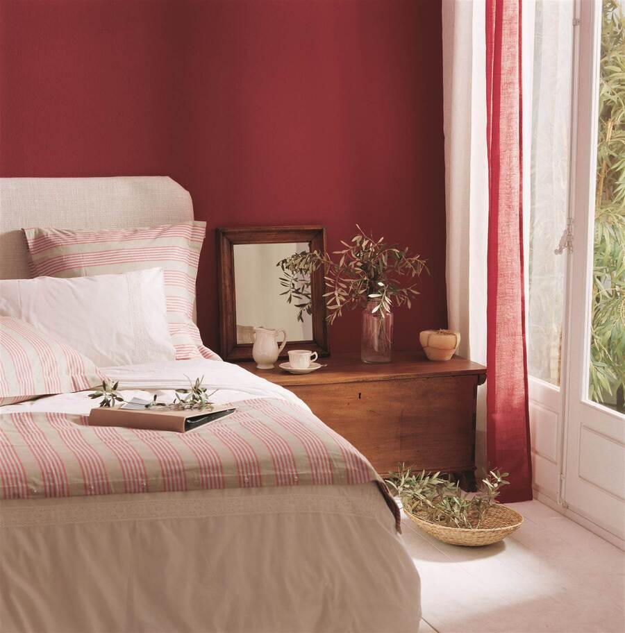 Dormitorio con una pared pintada magenta, cortinas de lino, también en magenta, y un velador de madera. A su lado está una cama con cobertor y almohadas blancas y una piecera y cojines color crema con líneas rosadas.