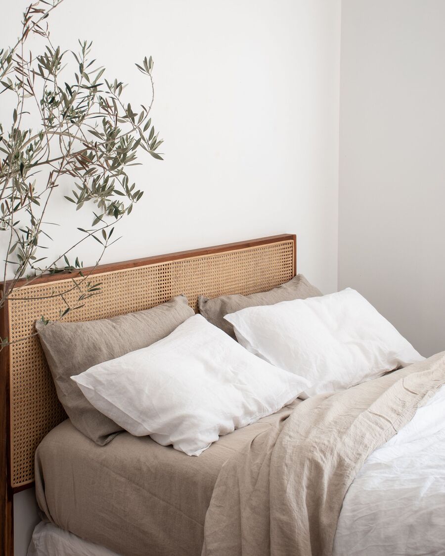 Detalle de una cama con respaldo de ratán, sábanas de lino color arena y almohadas blancas y funda de plumón blanca.