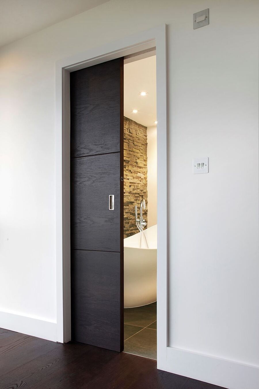 Pasillo de muros blancos y suelo de madera oscura con una puerta corredera de madera negra que da hacia un baño.