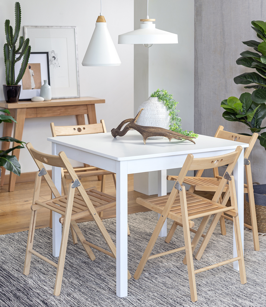 Comedor con una mesa cuadrada blanca y 4 sillas de madera plegables. Del techo cuelgan 2 lámparas blancas con detalles de madera.