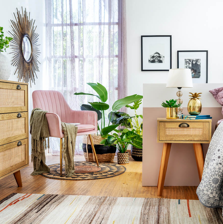 Dormitorio de estilo Boho Chic con 2 alfombras en tonos tierra, plantas dentro de canastos de fibras naturales y maceteros con diseños éticos, una silla rosada pastel, una cómoda y velador de madera con detalles de ratán.