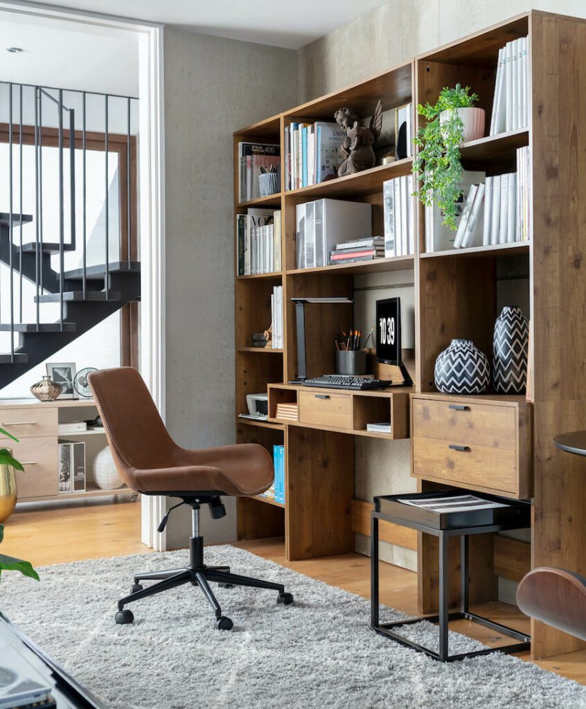 Zona de trabajo de estilo ejecutivo urbano con un gran mueble de madera, tipo biblioteca, con escritorio incorporado. Frente a él hay una silla de cuero café, que está sobre una gran alfombra gris con líneas blancas.