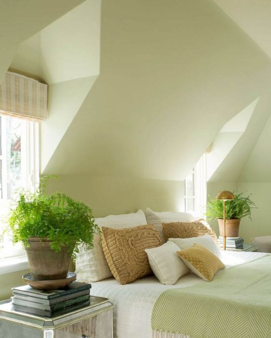 Dormitorio estilo campestre clásico con muro de color verde manzana. Cama de dos plazas con ropa de cama blanca y manta verde. Tiene 6 cojines en tonos blancos y mostazas. A los costados hay dos veladores espejados, con libros y plantas.