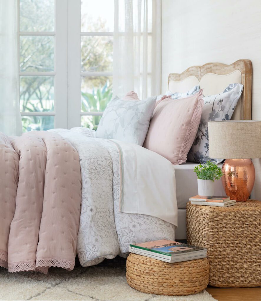 Dormitorio estilo clásico con una cama con respaldo de madera y acolchonado beige, frazadas rosa palo y blancos y almohadones color blanco, gris y rosa palo. Hay también veladores de mimbre con una lámpara con una pantalla beige y base de cobre. 