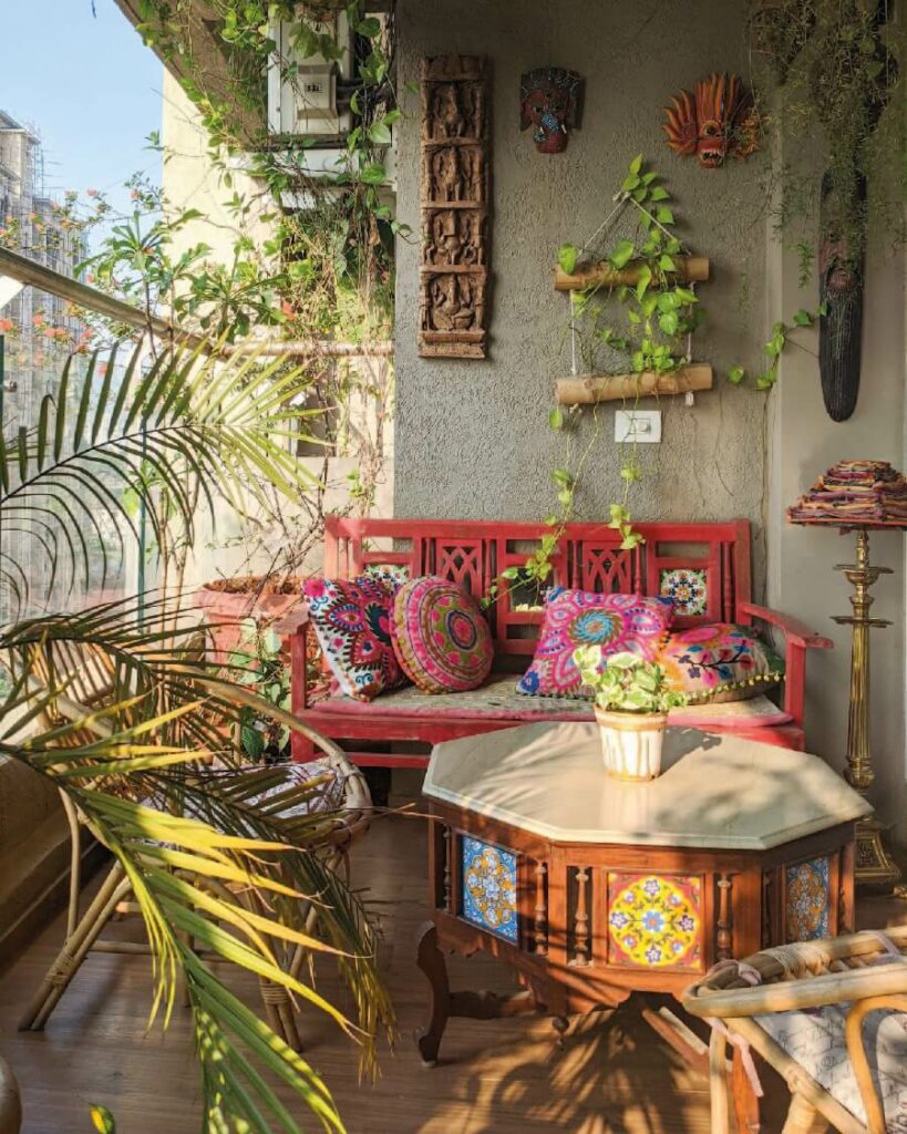 Terraza estilo boho chic con un banco rojo con varios cojines bordados, una mesa de centro octogonal estilo oriental y sillas de madera. Hay también varias plantas tropicales.