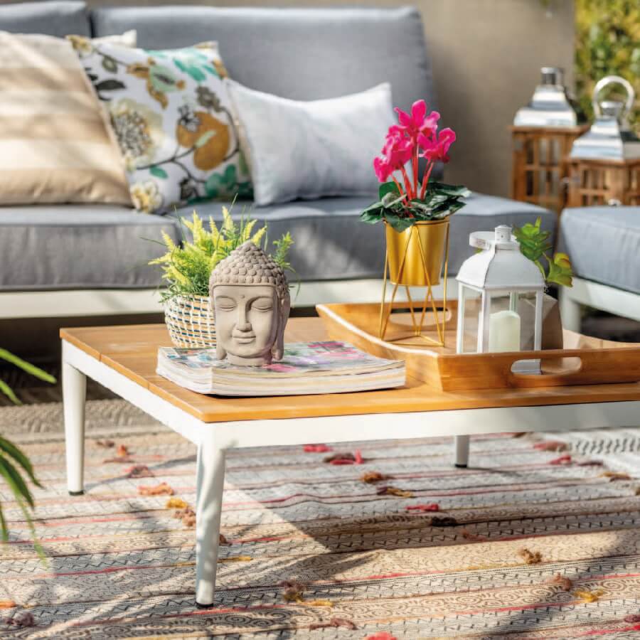Terraza con sofás grises con cojines de varios colores, con una mesa de centro de madera con base blanca. Sobre ella hay decoraciones de flores, velas y estatuilla de buda.