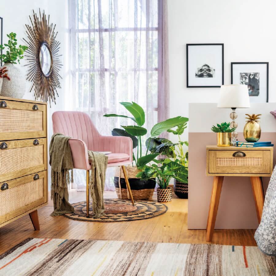 Habitación estilo boho chic, con una poltrona rosada con una manta color arena y sobre una alfombra de mimbre. Hay plantas tropicales, muebles de madera clara con detalles en ratán.