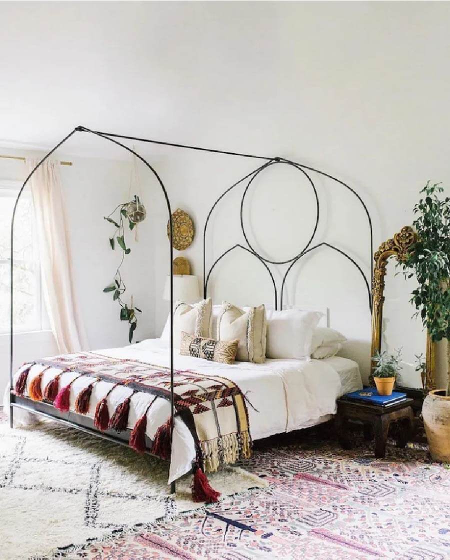 Dormitorio estilo boho chic con varias alfombras con iluestraciones, una cama con un marco negro alto con opatrones del medio oriente, con sábanas blancas y un pie de cama con ilustraciones étnicas.