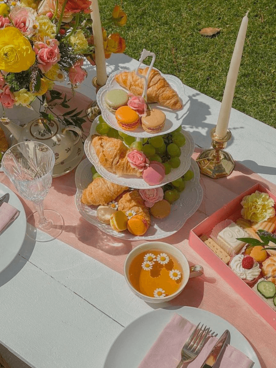 Picnic con un soporte para repostería de tres pisos con croissants y frutas, una caja de dulces, tazas de té, teteras, menaje y velas blancas.