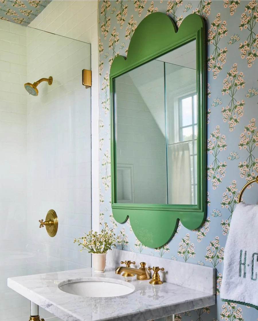 Baño estilo vintage, con papel mural de flores pequeñas en tonos celeste. Hay un espejo de marco verde, con formas curvas, lavamanos de mármol y ducha con grifería dorada.