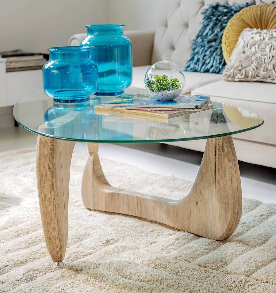 Mesa de centro de vidrio con patas de madera. Dos jarrones azules encima junto a revistas y un mini terrario. Alfombra y sofá color crema.