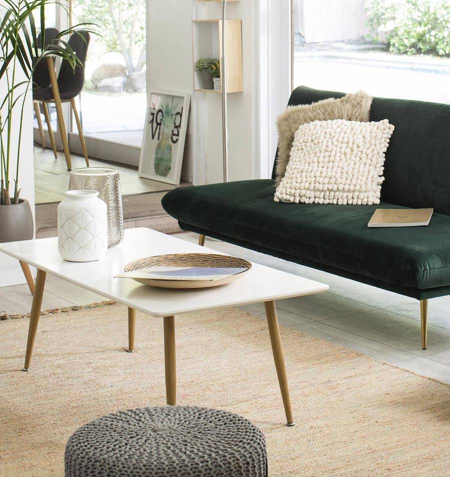 Mesa de centro blanco con patas largas de madera sobre una alfombra crema y junto a un futón verde oscuro y un pouf tejido gris.