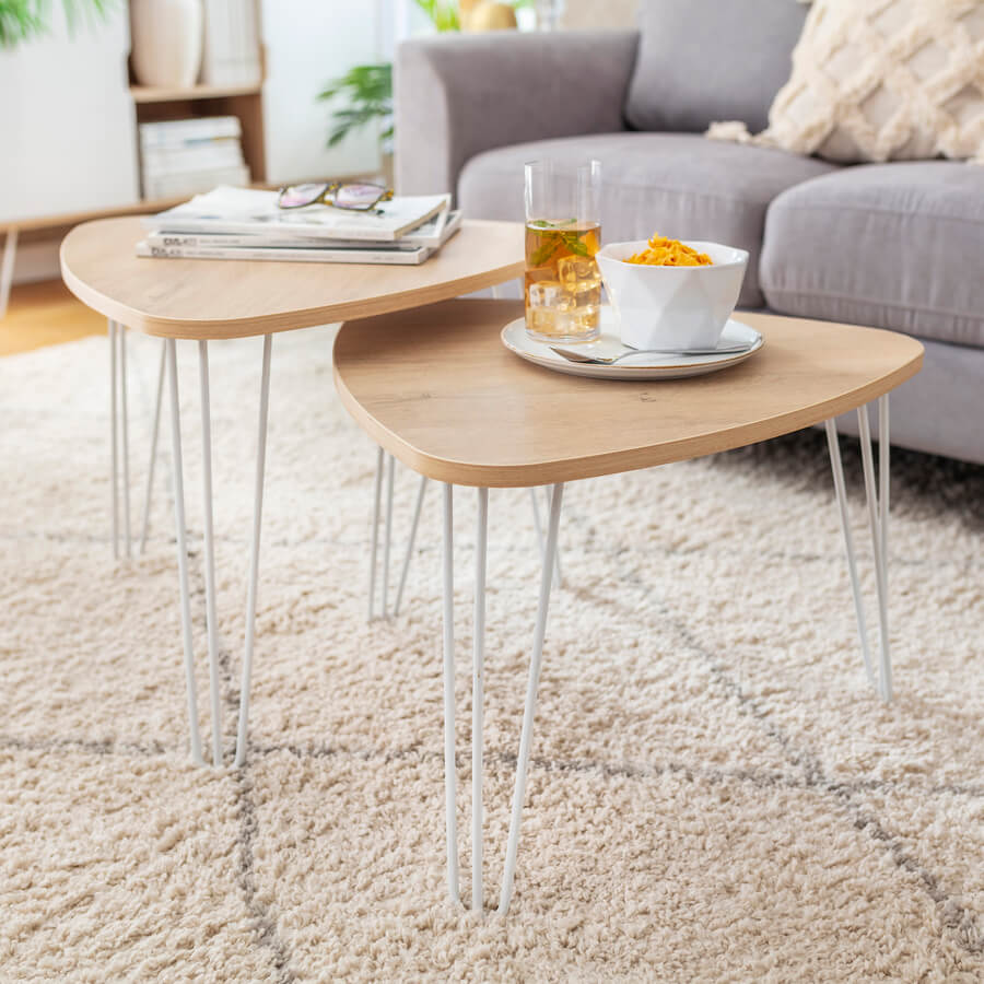 Set de dos mesas de centro, con base de madera clara y patas metálicas blancas. Alfombra decorativa color blanco con líneas grises, de estilo moderno o nórdico. Sofá de color gris claro.