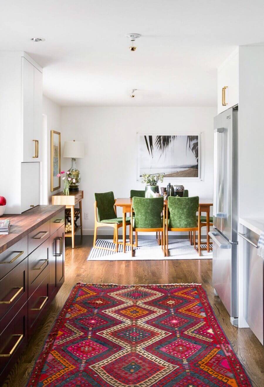 Comedor integrado con cocina. Muebles de cocina de madera oscura, alfombra decorativa estilo kilim en tonos burdeo, azul y naranjo. Comedor de madera anaranjada con sillas de tapiz verde.