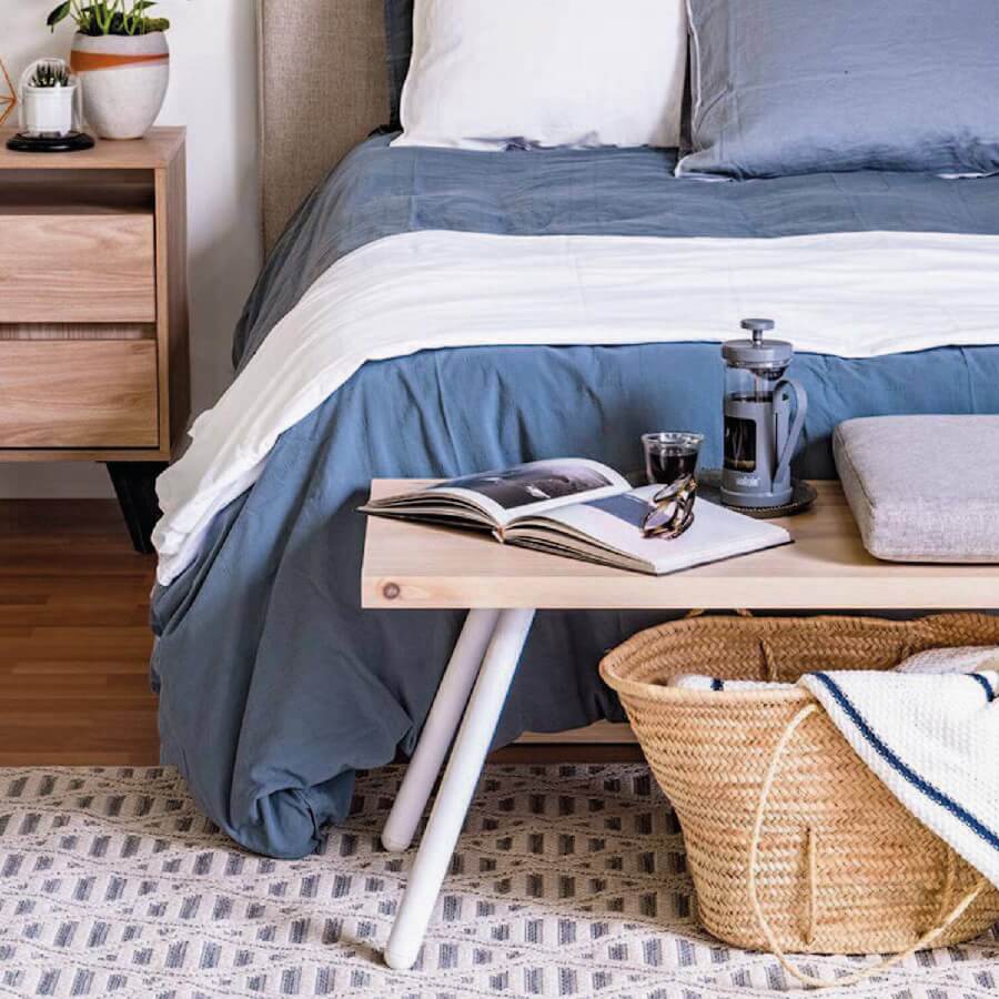 Banqueta de madera frente a una cama y sostiene un cojín, libros y cafetera. Bajo la banqueta hay una cesta de mimbre con una manta blanca.