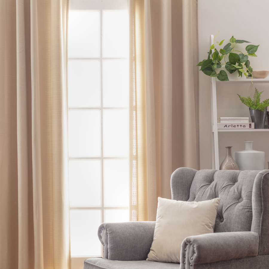 Rincón living moderno, con ventana y cortinas beige lisas. Sillón de un cuerpo gris estilo clásico, con cojín color blanco. Repisa blanca de varios niveles con libros y plantas.