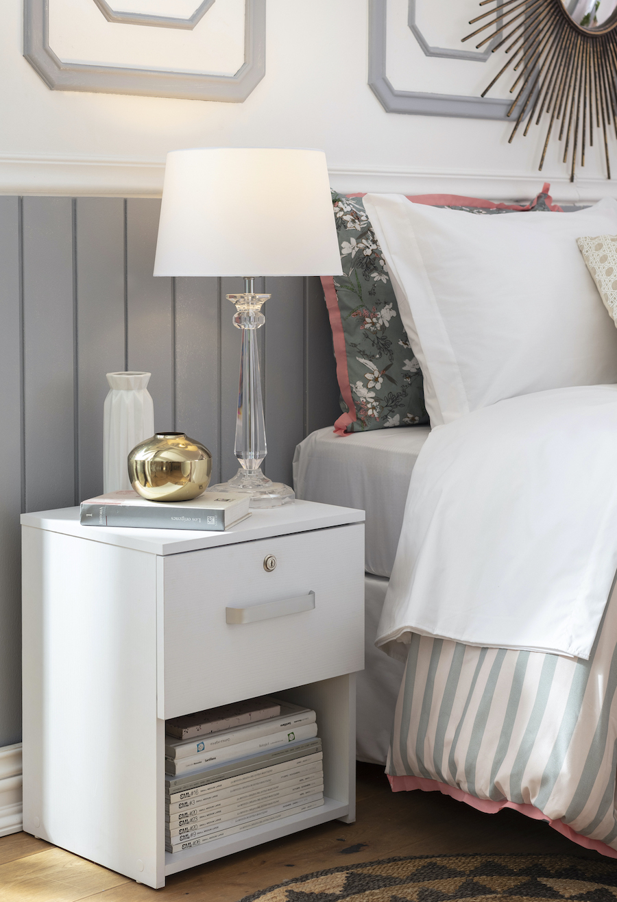 Lámpara con pantalla blanca y base transparente sobre un velador blanco que está juto a una cama con ropa de cama de tonalidades blancas y azules.