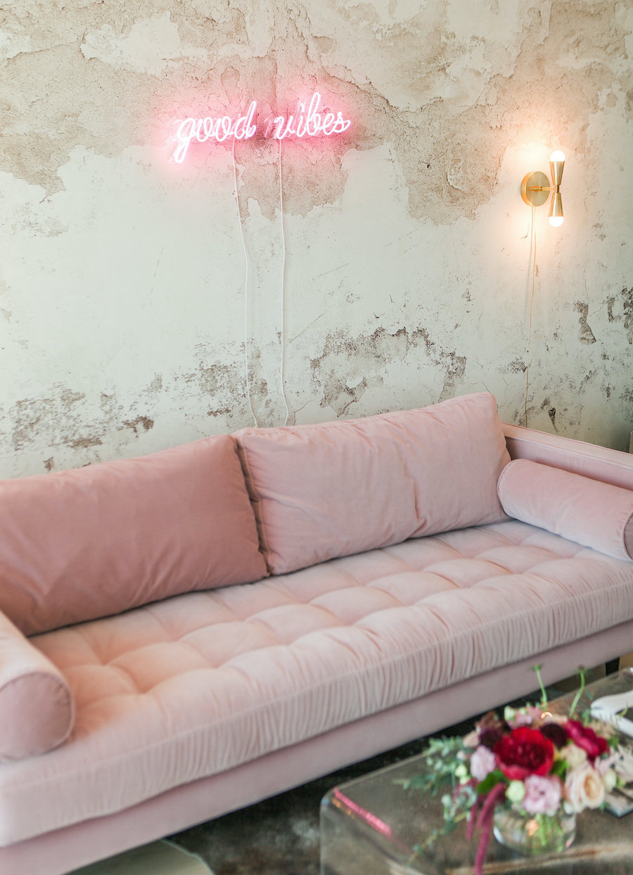 Luz de neón que dice "good vibes" sobre un sofá rosa palo. También hay un apliqué de pared dorado.