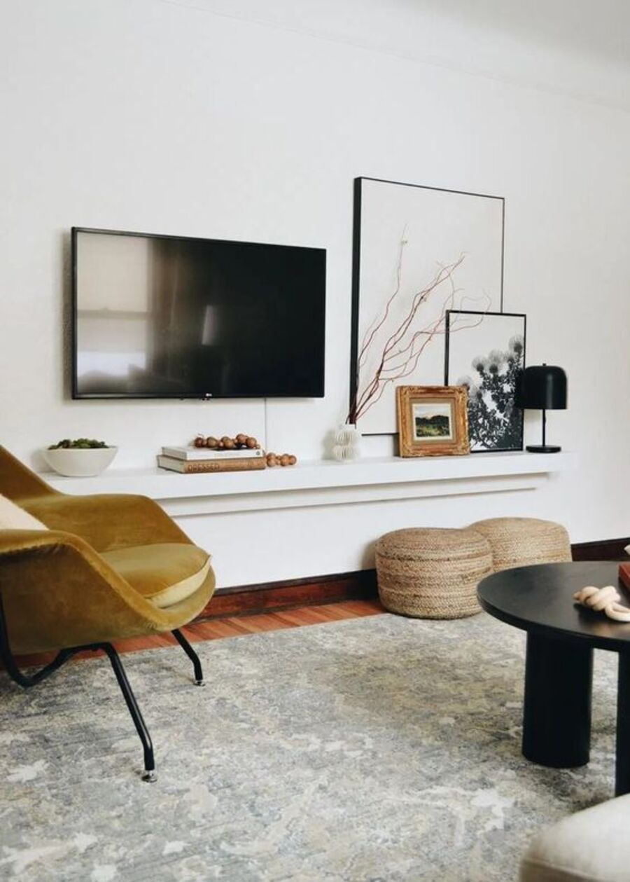 Living elegante y minimal, con repisa flotante alargada, que sostiene tres cuadros de diferentes tamaños y marcos. Un televisor colgado de la pared blanca. Alfombra gris y un sillón de terciopelo color mostaza.