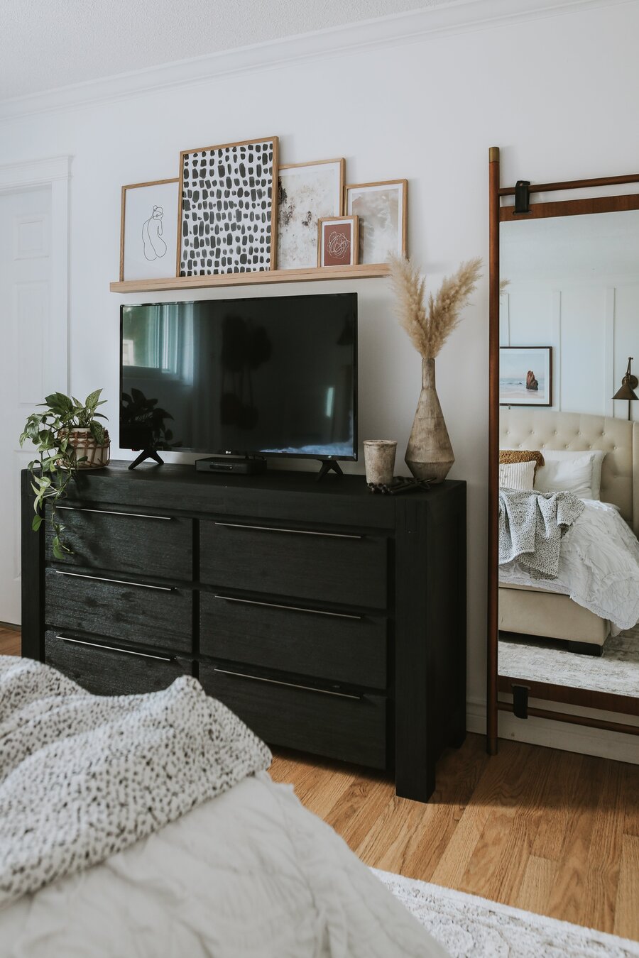 Dormitorio con cajonera de madera negra. Sobre ella el televisor y adornos con plantas. En el muro, una repisa flotante con varios cuadros apoyados, de diferentes tamaños. Al costado, un gran espejo con marco de madera.