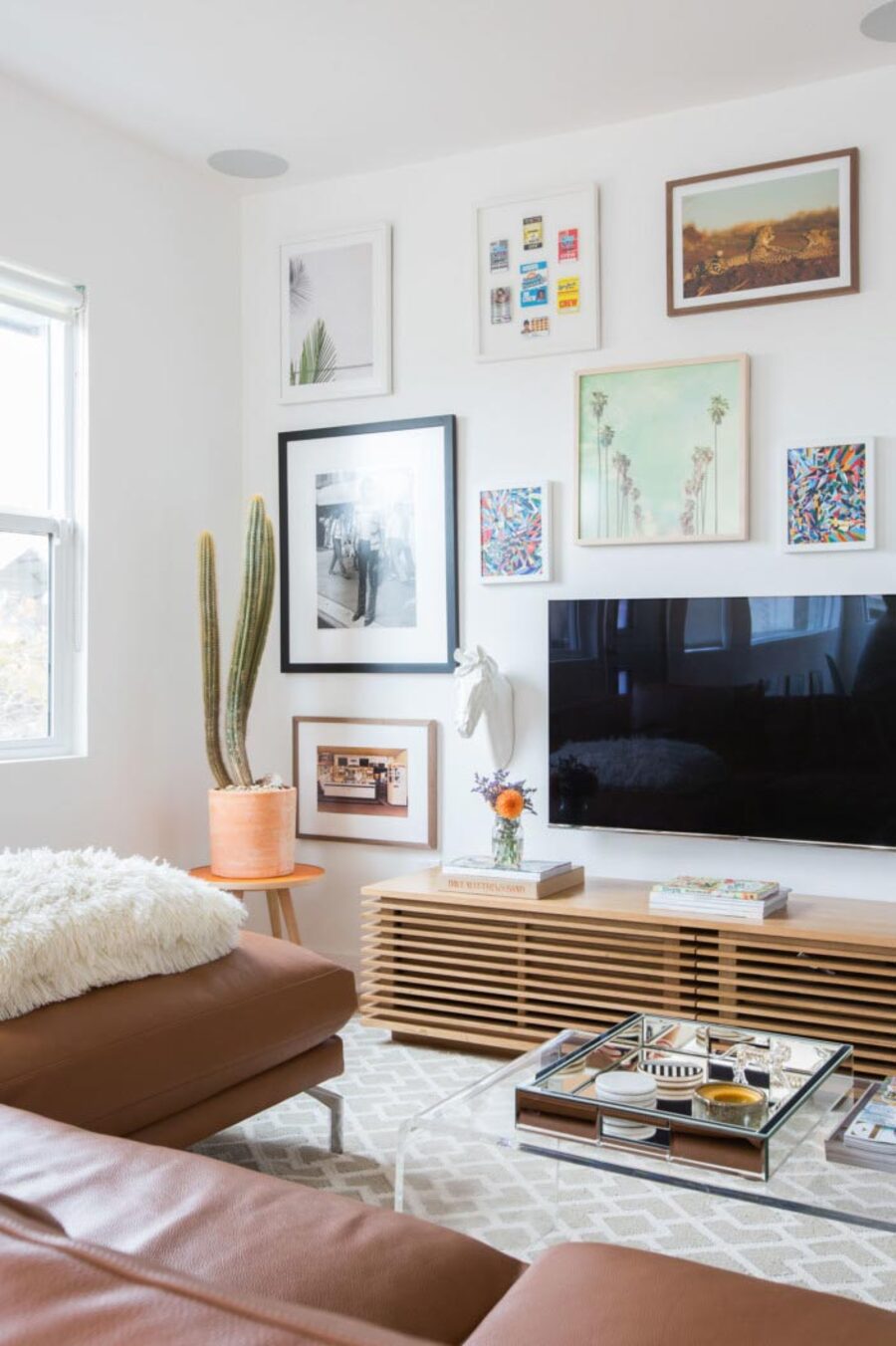 Muro galería con televisor colgado de la pared. Cuadros de diferentes formas, tamaños, colores y marcos. Mueble bajo de madera, mesa de centro transparente, sofá café y alfombra gris con blanco.