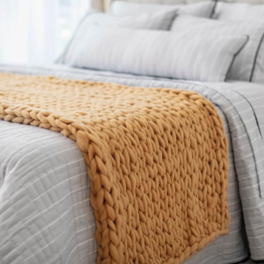Piecera tejida color mostaza sobre una cama con ropa de cama gris.