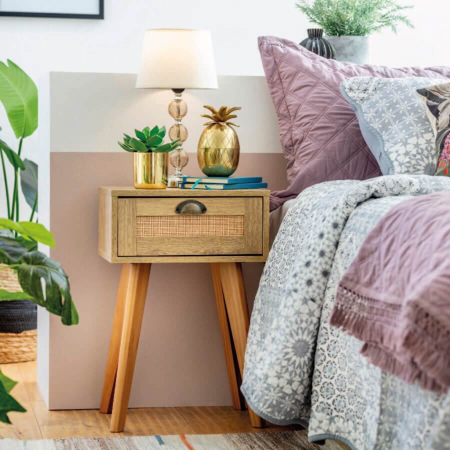 Velador de madera con detalles de mimbre que sostiene una lámpara con pantalla blanca, una maceta dorada con suculenta, cuadernos y un adorno dorado en forma de piña. Está al lado de una cama con ropa de cama de tonos rosa palo y gris.