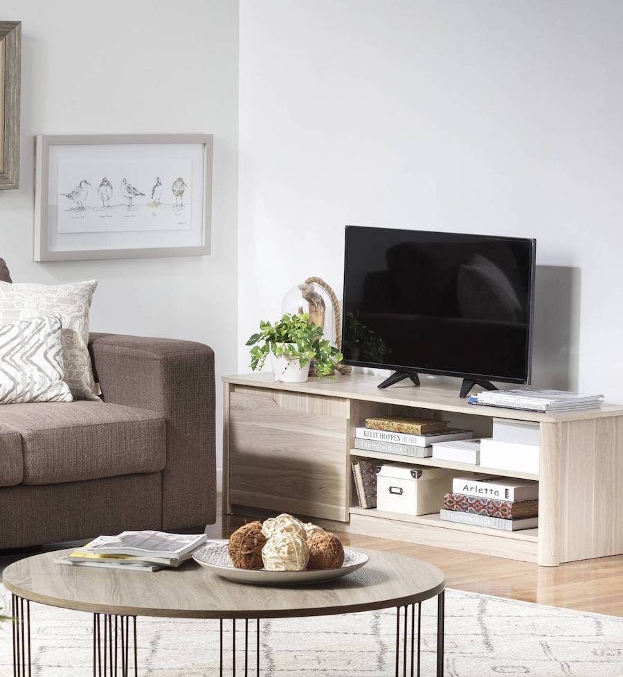 Living con un sofá café, mesa de centro redonda de madera, alfombra blanca y un mueble de tv de madera clara con un tv, macetas, libros y adornos.