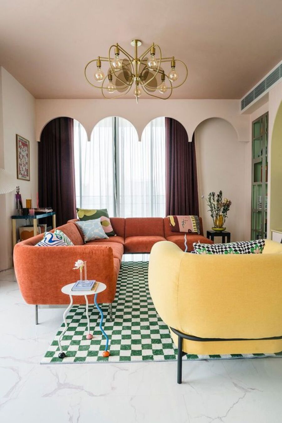 Living de estilo mid century, con piso blanco y alfombra de damero o patrón de ajedrez blanco y verde. Sillón en L de color naranjo, con cuatro cojines estampados. Sofá amarillo, lámpara colgante dorada.