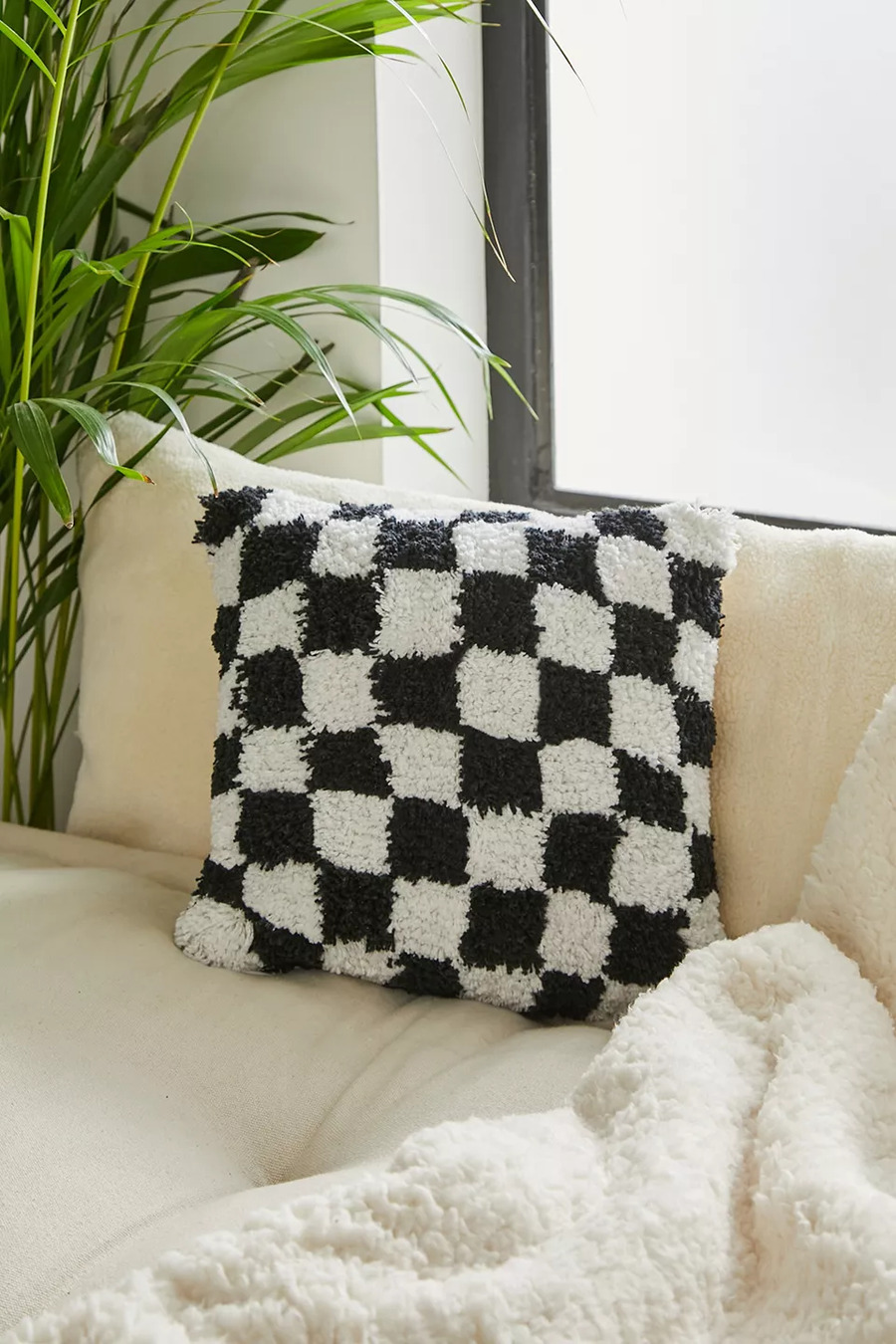 Idea de decoración con cojín de tela peluda, con patrón de ajedrez o damero en blanco y negro, sobre sofá color beige y manta de polar blanca.